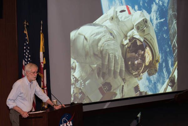 photo of Compton Tucker speaking at a podium in front of a large photo of Piers on a spacewalk