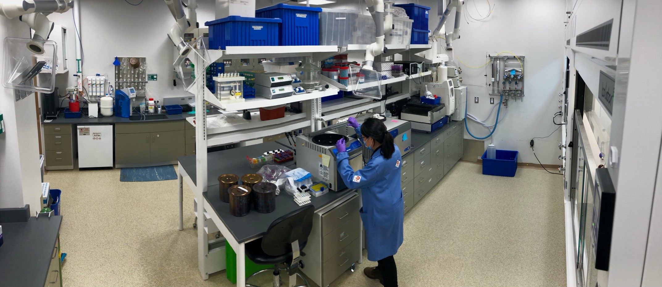 Woman in a blue lab coat working with a machine sat on a table with shelves behind that reach to the ceiling.